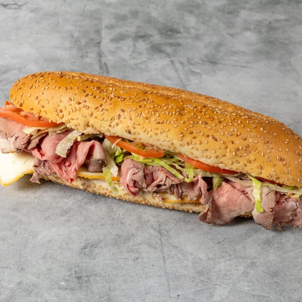 Long sesame-seed sub sandwich filled with roast beef, ham, cheese, lettuce, and tomato on a gray surface.