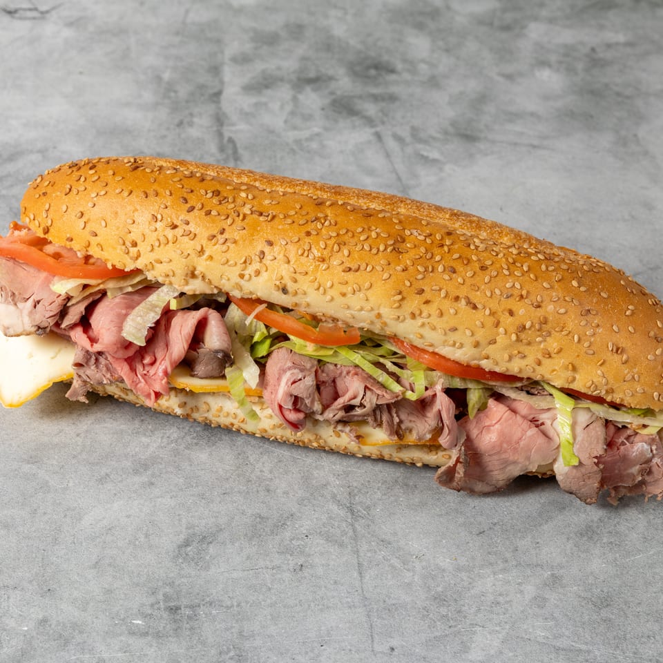 Long sesame-seed sub sandwich filled with roast beef, ham, cheese, lettuce, and tomato on a gray surface.