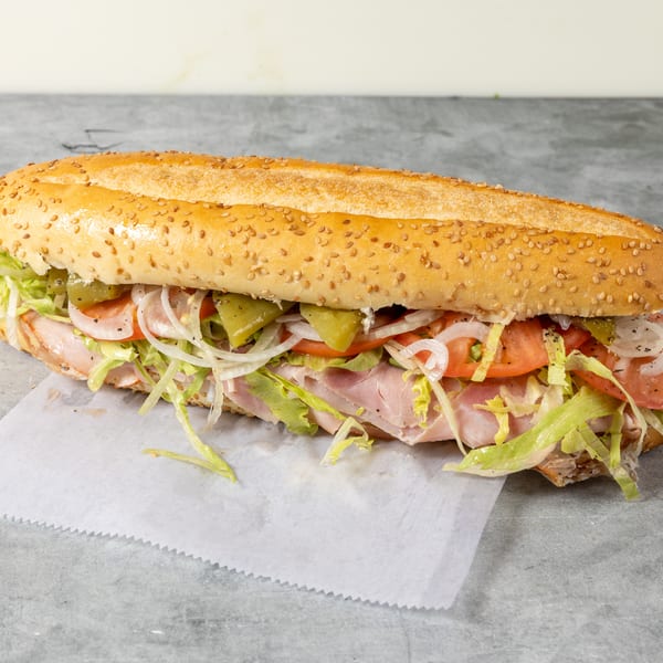 Sesame-seed sub sandwich filled with sliced ham, lettuce, tomatoes, onions, and green peppers on wax paper.