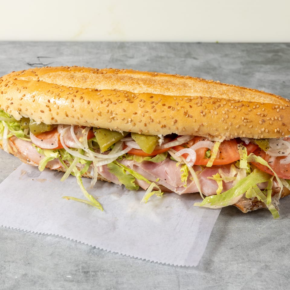 Sesame-seed sub sandwich filled with sliced ham, lettuce, tomatoes, onions, and green peppers on wax paper.