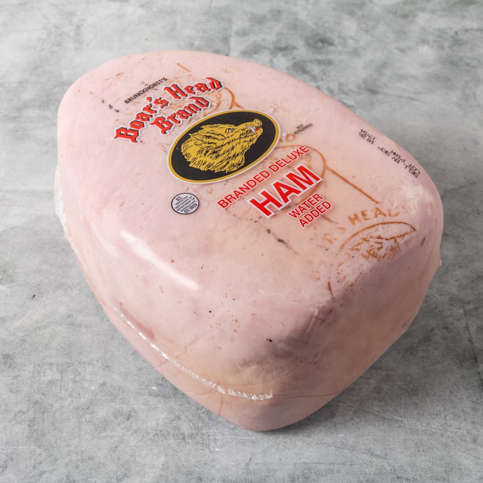 Large packaged pink ham labeled Rogerwood Farm, branded deluxe ham with added water, on a gray surface.