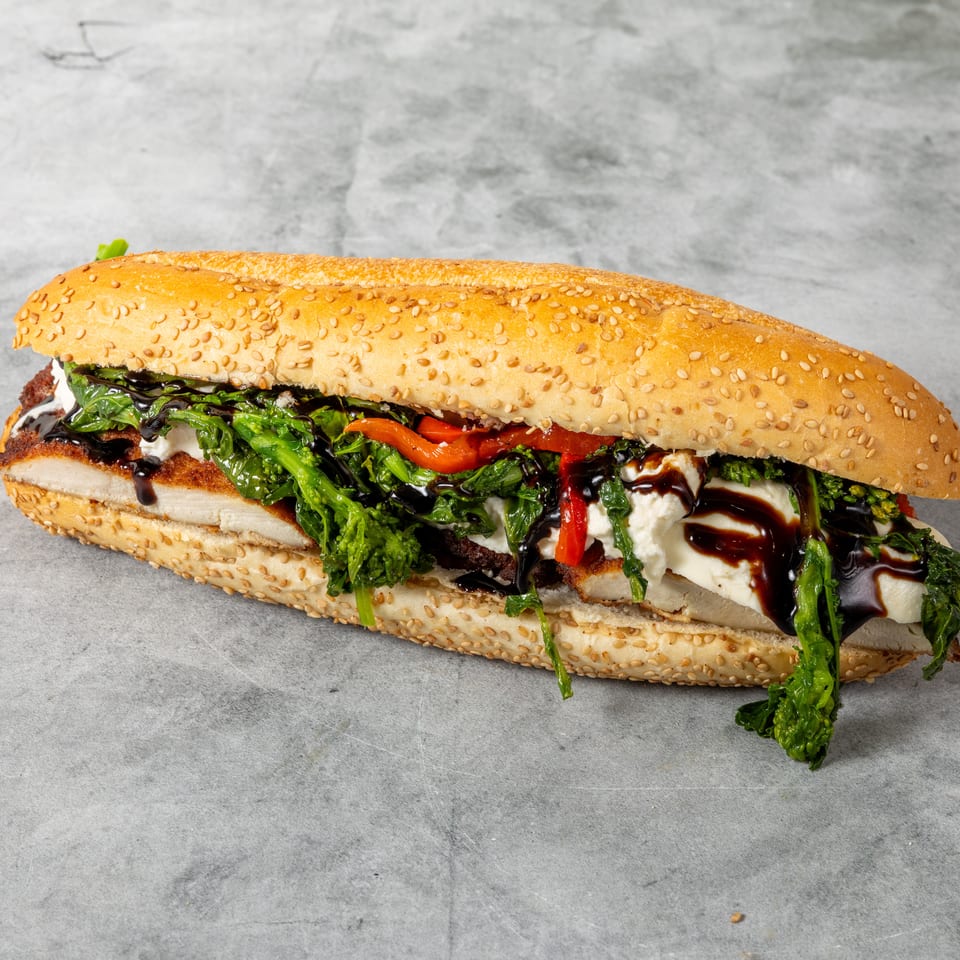 A long sesame seed hero sandwich filled with chicken cutlet, broccoli rabe, roasted peppers, and fresh mozzarella, drizzled with balsamic glaze.