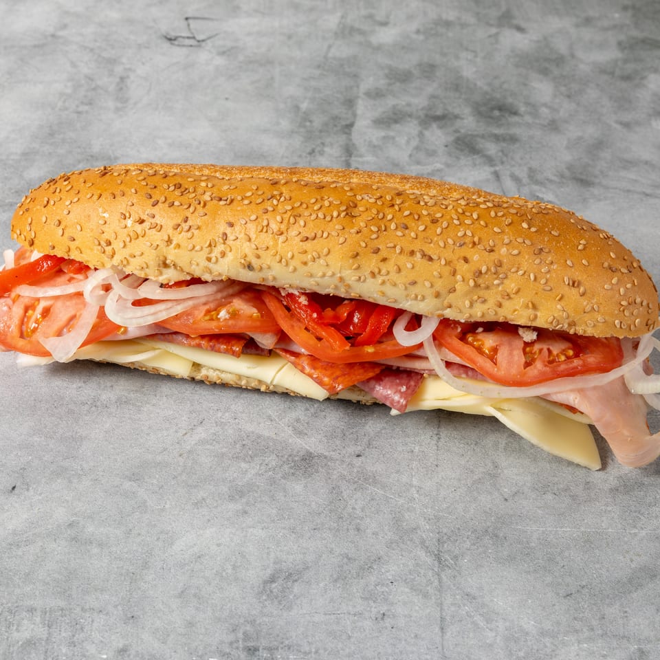 Sesame-seed sub sandwich filled with sliced meats, cheese, tomatoes, onions, and red peppers on a gray surface.